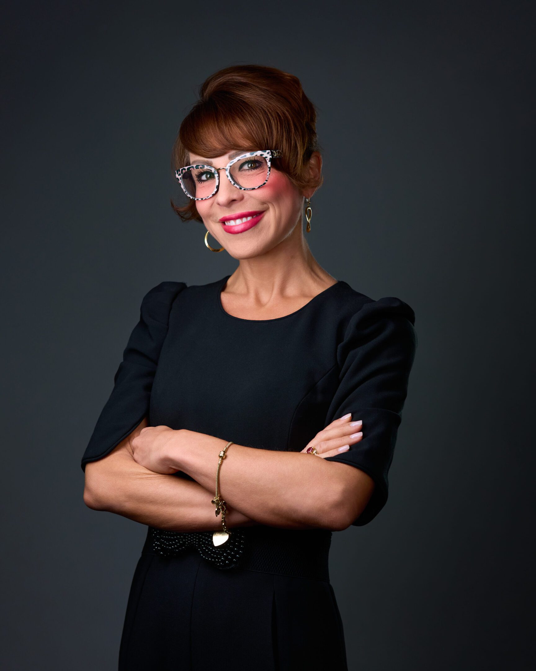 Confident woman in glasses and black dress smiles with arms crossed.